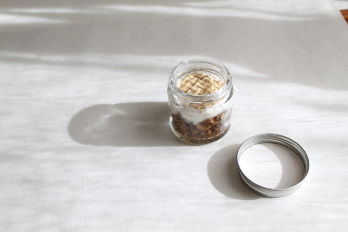 A small glass jar with a white screw-on lid labeled "MADISON GREEN - ROSE AND REFRESH MORNING AROMATHERAPY INHALER," containing mixed dried botanicals for natural stress relief, sits against a plain white background.