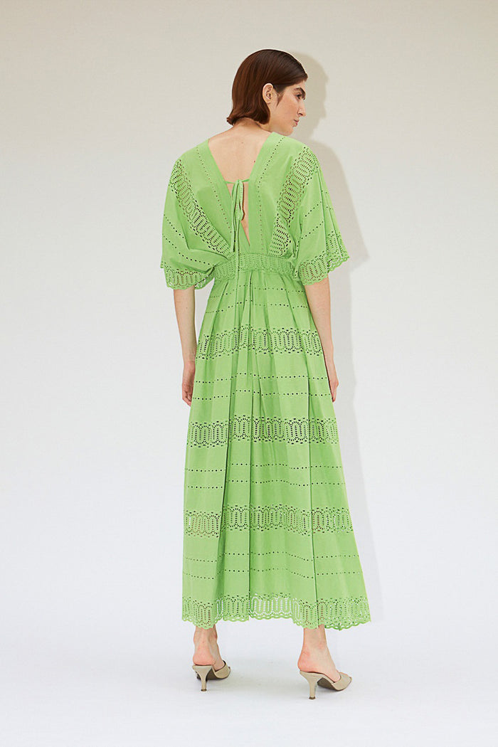 A person wears the MILVA SANGALLO DRESS, a light green cotton v-neck with eyelet details, short wide sleeves, and an ankle-length hem, standing against a plain white textured background.