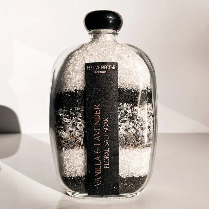A clear glass bottle of NATIVE NECTAR VANILLA & LAVENDER FLORAL SALT SOAK with black and white layers sits on a white surface, inviting you to a soothing plant-based lavender vanilla bath.