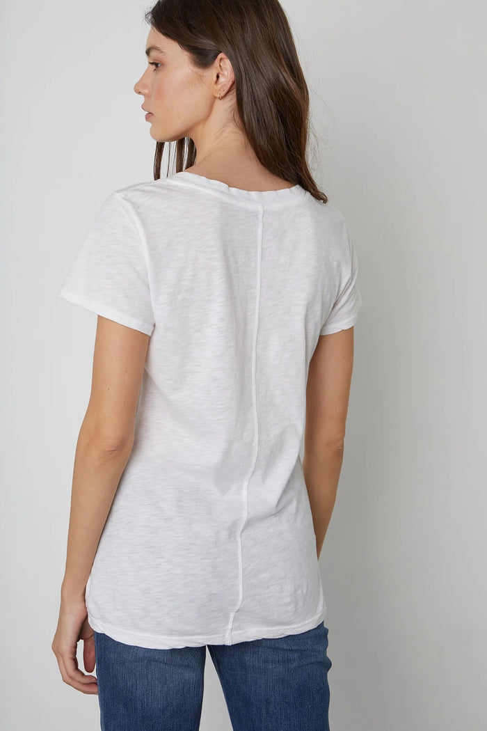 A woman with long brown hair wears the VELVET by Graham & Spencer Lilith V Neck Slub Cotton Tee and blue jeans, standing against a light gray background and looking at the camera with a neutral expression.