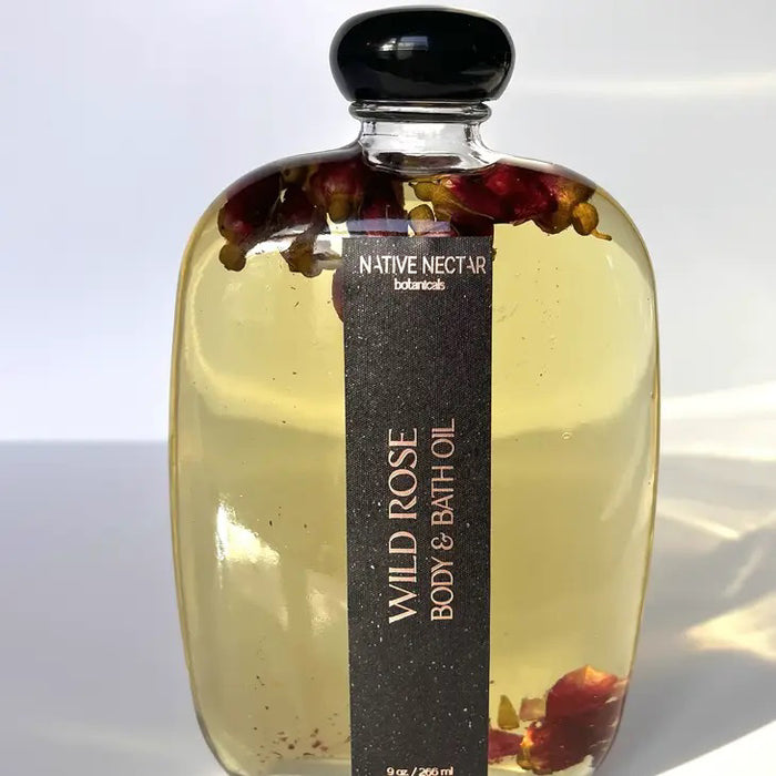 A clear bottle of NATIVE NECTAR WILD ROSE BODY OIL with rose petals and a black label highlights this botanical-infused moisturizing oil against a plain white background.