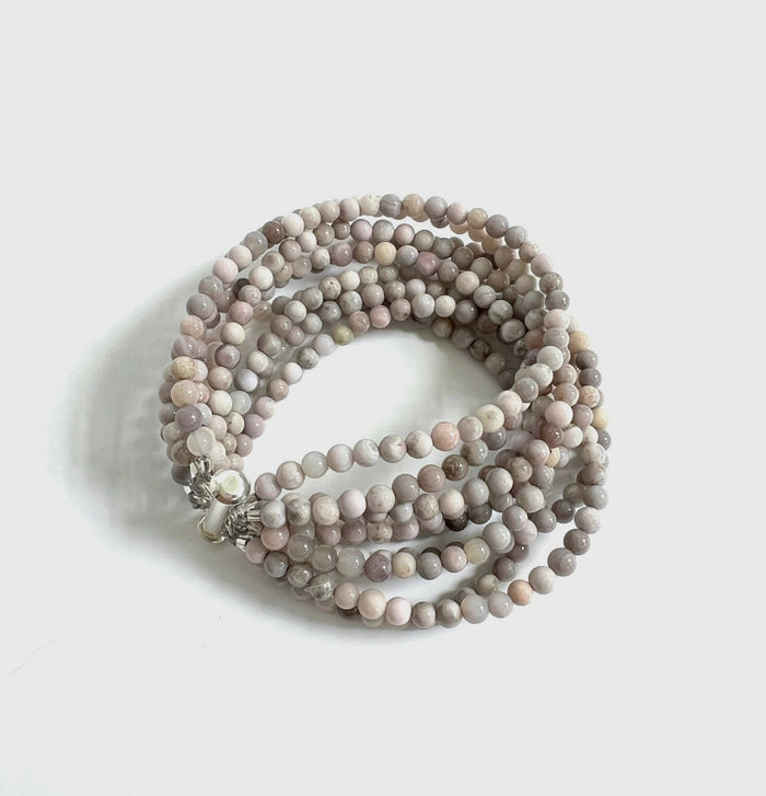 The ANN LIGHTFOOT - NINE STRAND SAKURA AGATE CUFF features smooth, round sakura agate beads in light grey and beige. Coiled with multi-strands, the bracelet has 4mm beads and is secured by a stainless steel clasp against a plain white backdrop.