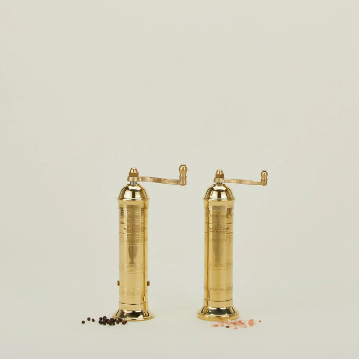 The ATLAS BRASS SALT MILL 9" by PEPPER MILL IMPORTS is a tall, elegant handcrafted Greek product with a cylindrical design and detailed engravings. It includes a top crank handle for grinding and boasts a sleek, polished finish against a white background.