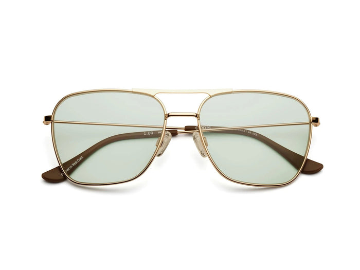 A man with a beard and glasses wears CADDIS - HOOPER READERS by CADDIS, featuring a lightweight, nickel-free metal alloy frame. He pairs them with a black cap and graphic tee, standing before vibrant embroidered art of faces and geometric patterns.