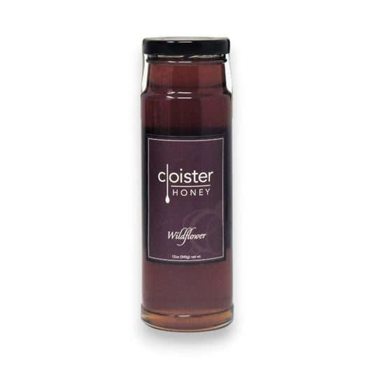 A glass jar of CLOISTER HONEY - HONEY WILDFLOWER 12OZ features a purple label with a black cap. This 12 oz (340g) wildflower honey offers rich, amber floral sweetness.