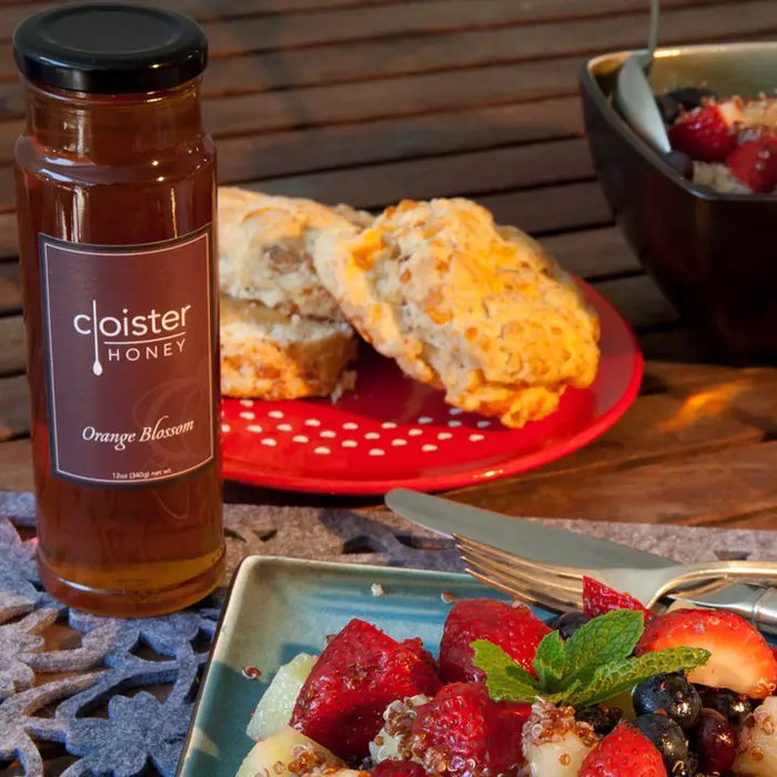 A bottle of CLOISTER HONEY - ORANGE BLOSSOM HONEY 12OZ by CLOISTER HONEY sits next to a red plate with biscuits, with fresh fruit and quinoa garnished with mint in front, and a bowl of mixed fruit on the wooden table.