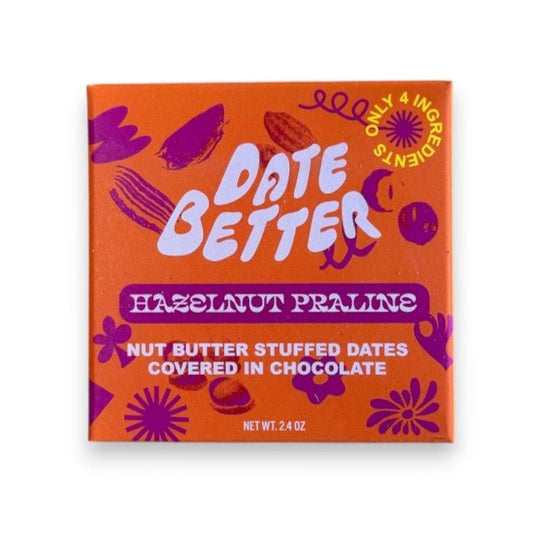 The "DATE BETTER - HAZELNUT PRALINE COVERED DATES" from DATE BETTER SNACKS features orange packaging with bold pink and purple text, adorned with abstract flower and leaf patterns. This 85% dark chocolate Medjool date treat weighs 1.8 oz.
