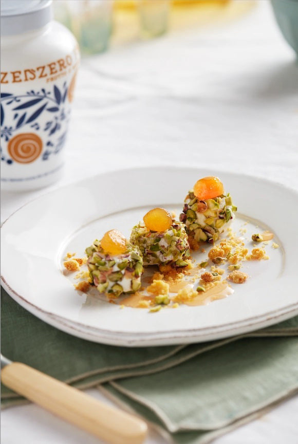 Three small, round desserts topped with orange garnish and chopped pistachios sit on a white plate, drizzled with FABBRI - CANDIED GINGER IN SYRUP by GOURMET FOOD SOLUTIONS. Crumbled toppings surround them; a jar rests blurred in the background.