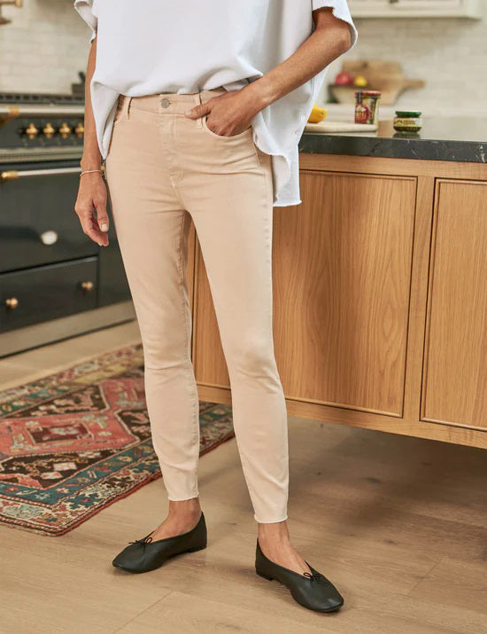 A person stands in a kitchen, showing only their lower half, wearing FRANK & EILEEN’s SLIGO SKINNY JEAN in light beige with a white top and black flats, set against a stove, countertop, and patterned rug.