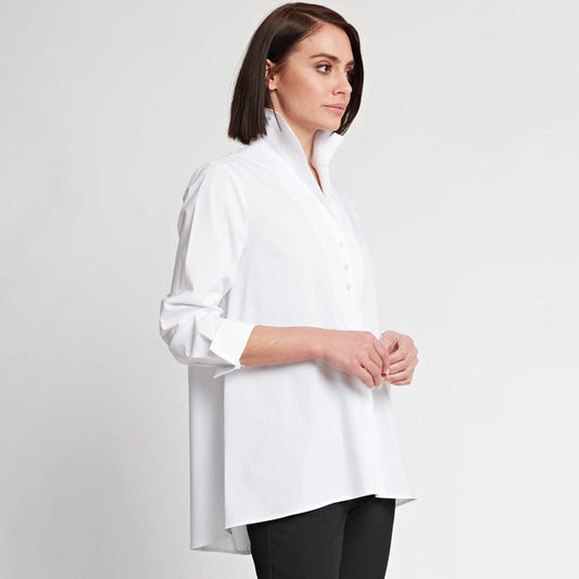 A woman with straight brown hair models the HINSON WU BETTY LONG SLEEVE TUNIC, a loose-fitting, white, A-line button-up shirt with a standing collar and rolled sleeves, paired with black pants against a plain light background.