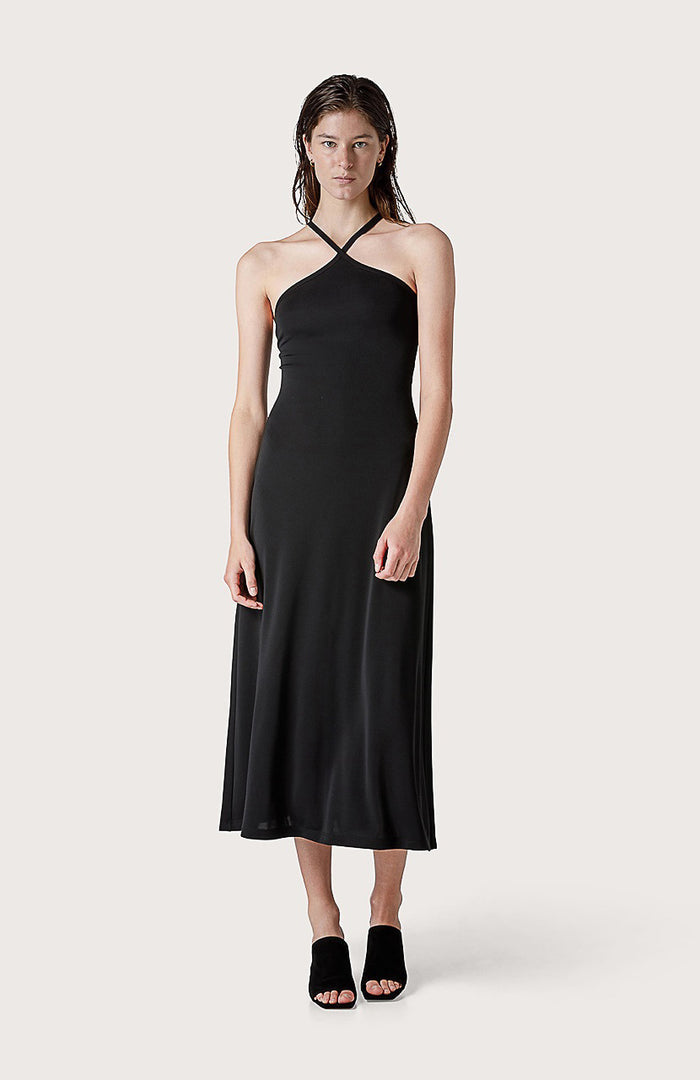 A woman faces forward in the SEVENTY JERSEY DRESS, a sleeveless black style with crisscross neckline straps, paired with black open-toe heeled sandals—ideal for summer parties—set against a plain light background.