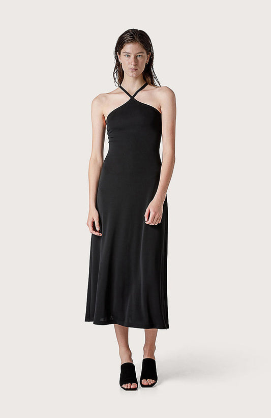A woman faces forward in the SEVENTY JERSEY DRESS, a sleeveless black style with crisscross neckline straps, paired with black open-toe heeled sandals—ideal for summer parties—set against a plain light background.