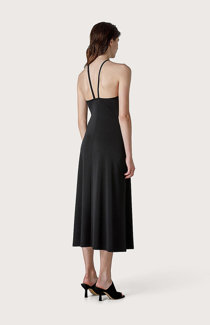 A woman faces forward in the SEVENTY JERSEY DRESS, a sleeveless black style with crisscross neckline straps, paired with black open-toe heeled sandals—ideal for summer parties—set against a plain light background.