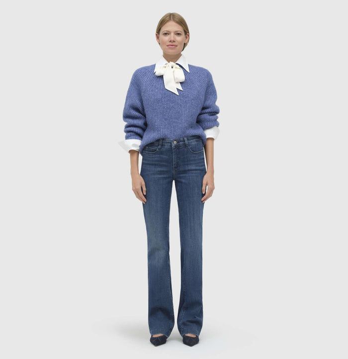 A person stands wearing a blue sweater, white shirt with a bow, and MAC's DREAM BOOT AUTHENTIC DENIM jeans. They have short light brown hair and pose against a plain light gray background.