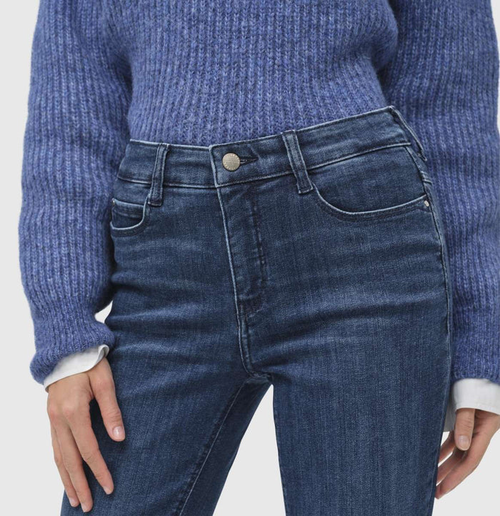 A person stands wearing a blue sweater, white shirt with a bow, and MAC's DREAM BOOT AUTHENTIC DENIM jeans. They have short light brown hair and pose against a plain light gray background.