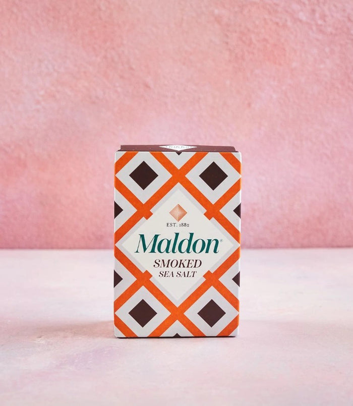 A box of GOURMET FOOD SOLUTIONS MALDON - SMOKED SEA SALT with an orange, brown, and white geometric design sits on a light pink surface against a textured pink background.