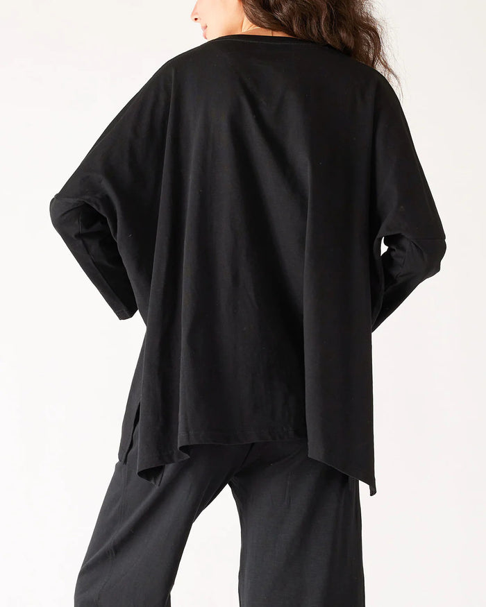A woman with wavy brown hair models the MERSEA - CATALINA SLUB TEE—an oversized, long-sleeved white top with thin gray stripes—by MERSEA. She pairs it with blue jeans and stands against a plain, light background, hands behind her back.