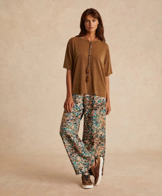A woman with long brown hair wears the MOMONI - IORA LUREX JERSEY T SHIRT in gold, paired with beige and blue wide-leg floral pants and brown sneakers, standing against a neutral studio background.