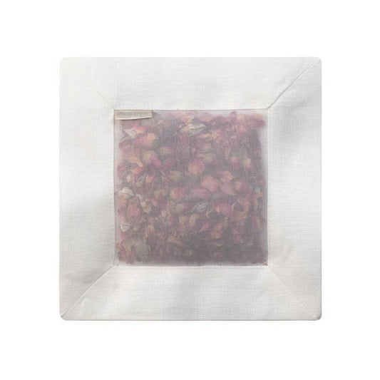 The ROSE SQUARE SACHET IVORY LINEN by E WIGHTMAN & CO is a white linen sachet with a see-through center, revealing delicate pink rose petals. The square sachet is shown on a plain white background, emphasizing its elegant contents.