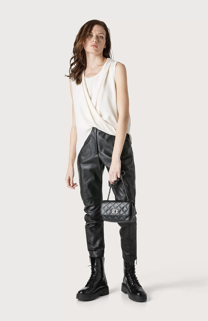 A woman stands against a plain background wearing the SEVENTY Jersey Sleeveless with Draped Hemline top, black leather pants, black lace-up combat boots, and holding a small black quilted handbag.