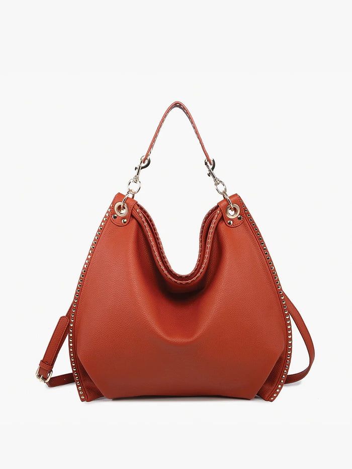 The STUDDED SLOUCHY HOBO by JEN & CO is a brown leather handbag with a slouchy design, featuring a shoulder strap and metal ring accents on the handle attachments. It has a smooth texture and structured shape against a white background.