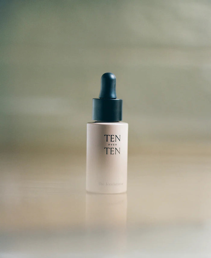 A glass dropper dispenses fast-acting serum into a pink bottle labeled "TENOVERTEN - THE ACCELERATOR QUICK DRY" by TEN OVER TEN, with a matching box beside it on a textured surface.