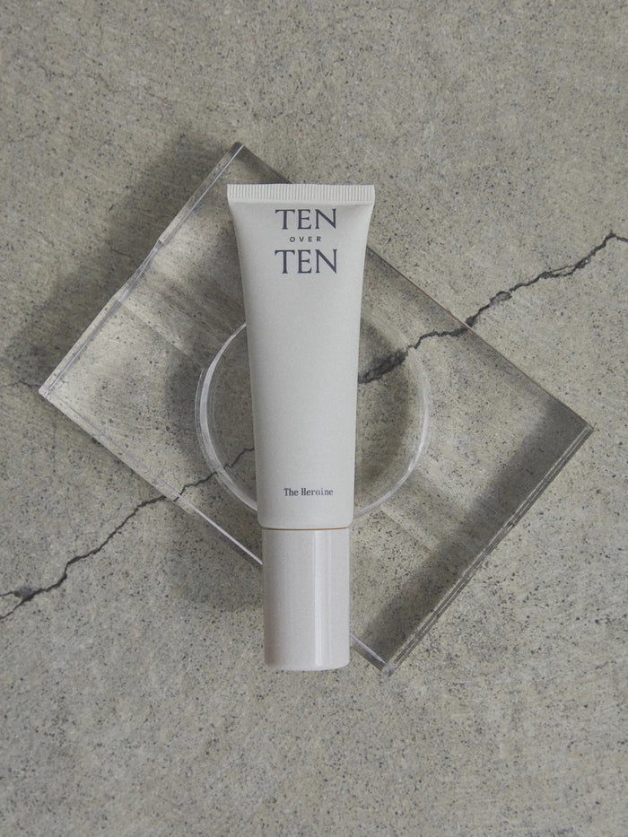 A beige tube labeled "TENOVERTEN - THE HEROINE HAND LOTION" by TEN OVER TEN rests on a clear rectangular platform atop a textured, cracked concrete surface.