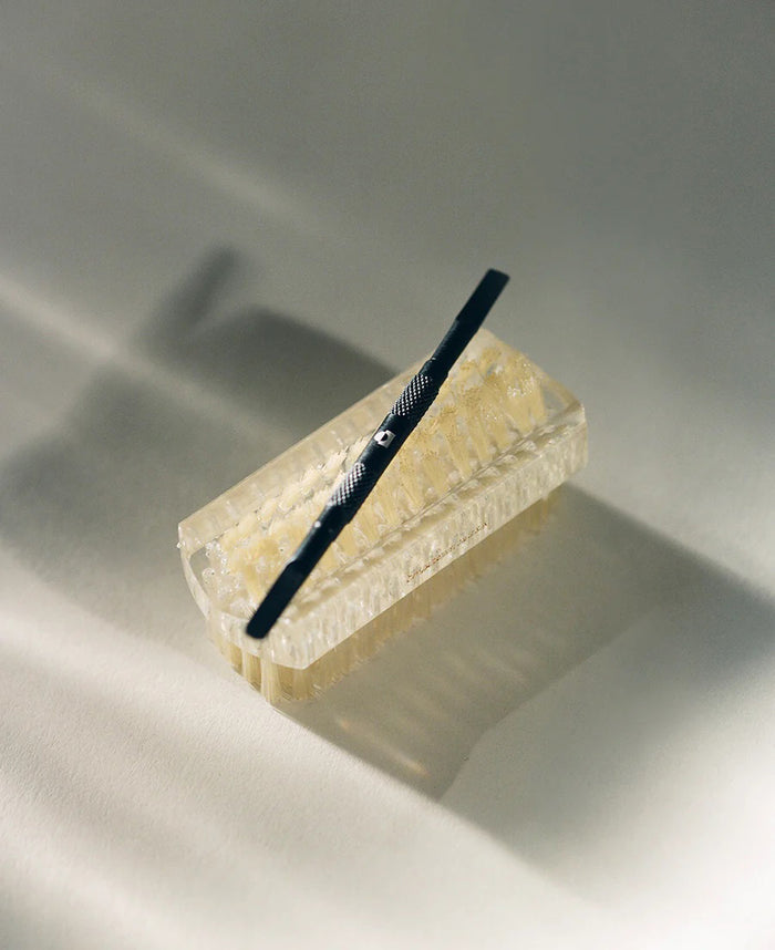 A black metal razor sits diagonally on a translucent, textured bar of soap next to TEN OVER TEN’s THE PUSH BACK CUTICLE tool, both casting soft shadows on a light surface.