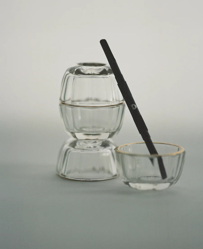A black metal razor sits diagonally on a translucent, textured bar of soap next to TEN OVER TEN’s THE PUSH BACK CUTICLE tool, both casting soft shadows on a light surface.