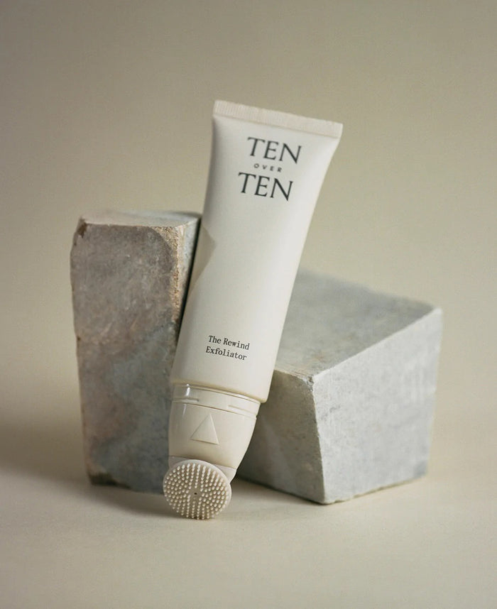 A tube of TENOVERTEN - THE REWIND HAND EXFOLIATOR by TEN OVER TEN, a gentle hand exfoliant with a textured applicator tip, is propped against two gray stone blocks on a neutral background.