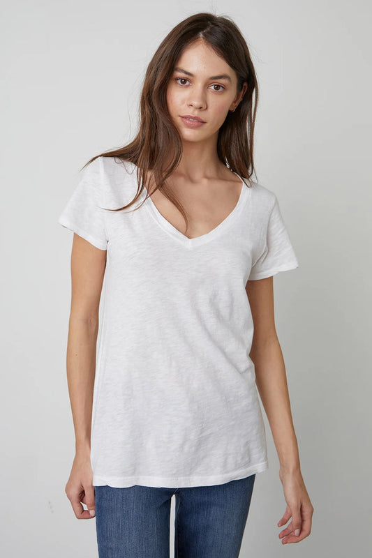 A woman with long brown hair wears the VELVET by Graham & Spencer Lilith V Neck Slub Cotton Tee and blue jeans, standing against a light gray background and looking at the camera with a neutral expression.